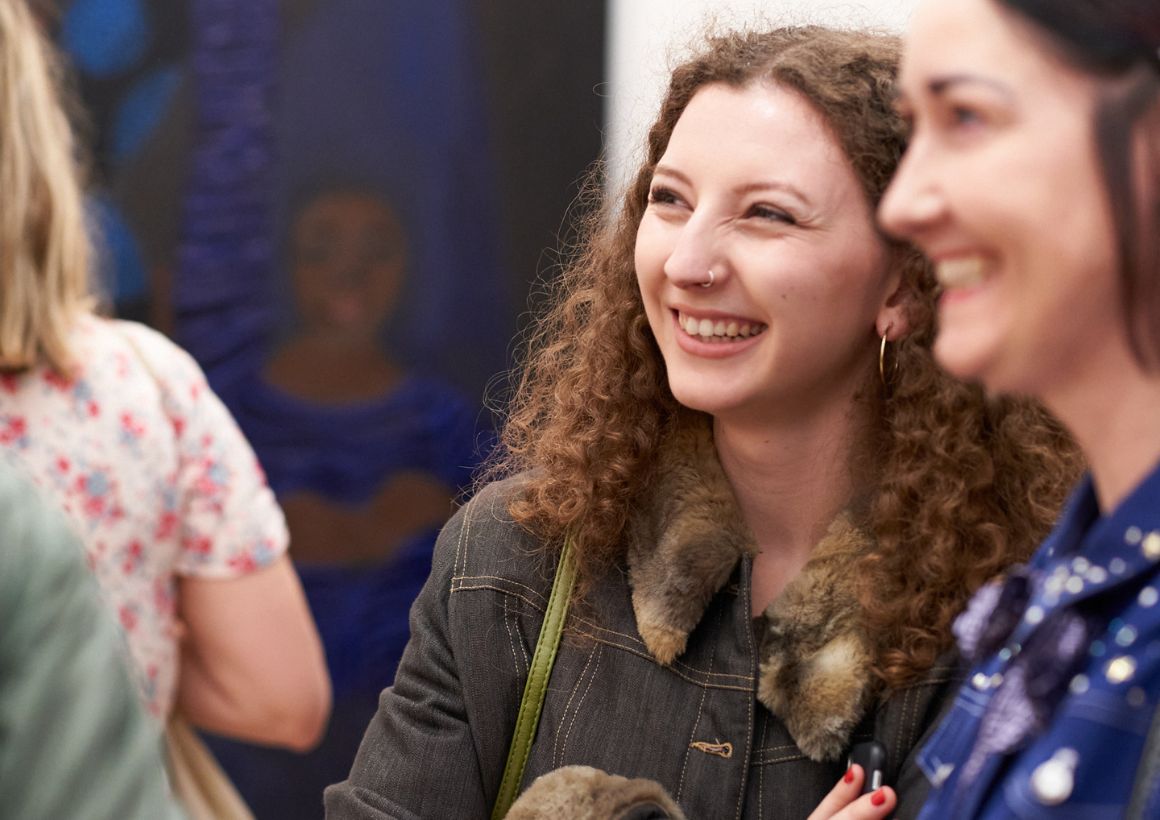 Two women smiling broadly among a crowd in an art gallery, with a painting on a white wall in the background