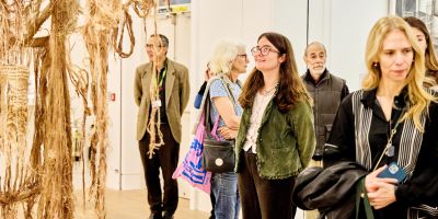 People gathered in a white walled gallery looking at the artworks. In the centre is a person with glasses wearing a green jacket looking up at a textile sculpture.