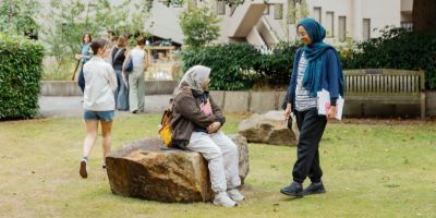 A grassed outdoor space with two large rocks in the foreground and a bench set further back. A woman is sitting on the nearest rock, looking up at another woman who is standing