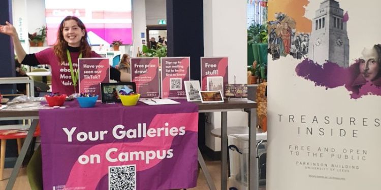 Cara, a white woman with red hair wearing a pink tshirt, stood behind a stall covered in Galleries merch and a purple tablecloth with white writing which reads "Your Galleries on Campus"