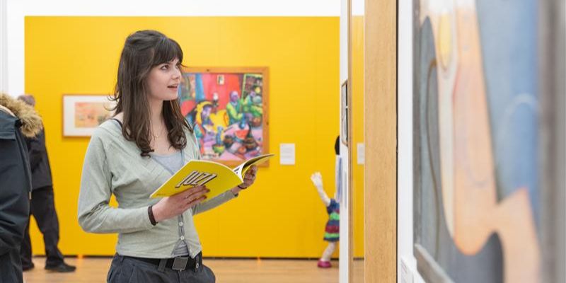 A person with long black hair wearing a pale green cardigan and holding a yellow book stands in a white-walled gallery looking at colourful paintings on the wall. Behind them is a wall painted yellow, more colourful artworks, and more people.