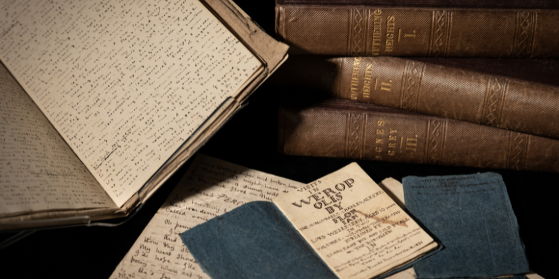 A selection of books and letters on black background, including a stack of three brown volumes, an open book on a stand, and a tiny open book with a blue cover.