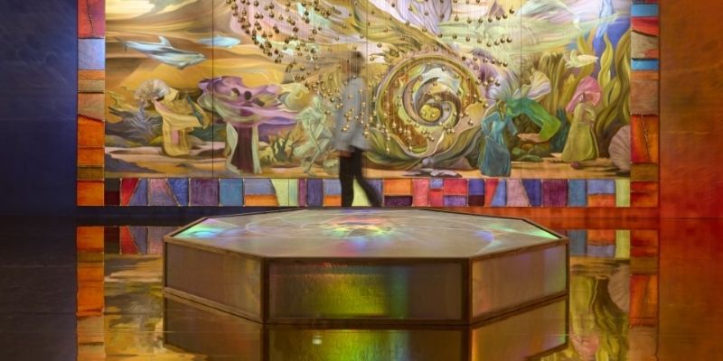 A colourful hexagonal block on a golden mirrored floor with golden spheres on threads hanging from the ceiling above it. In the background a person walks in front of a colourful multipaneled painting.