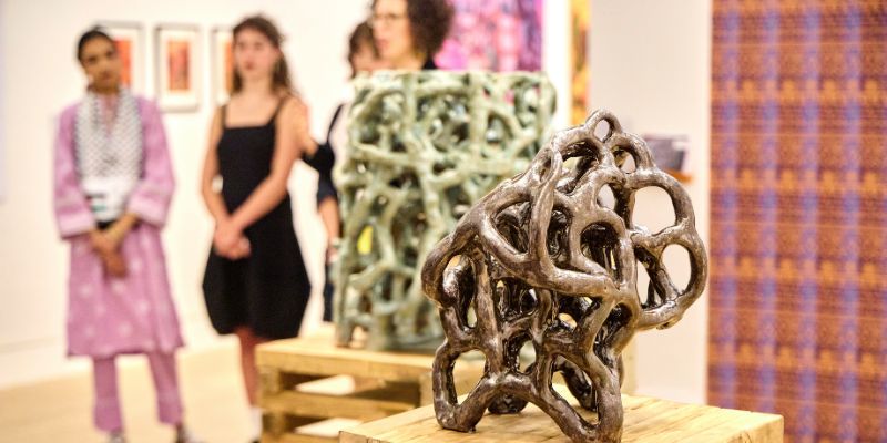 A woman wearing glasses and a headscarf looks at a work of art on a wall, surrounded by other gallery-goers