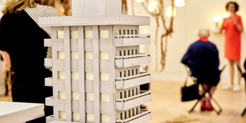 Close up of an architectural model of a block of flats. Behind it there are lights hanging from the ceiling, a large hessian sculpture, and a group of people listening to a talk.