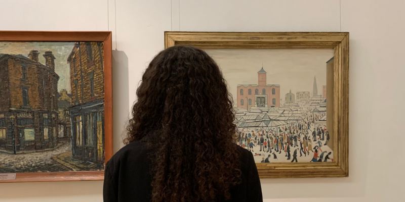 A photograph from behind of a person looking at Lowry's Market Stalls in The Stanley & Audrey Burton Gallery.