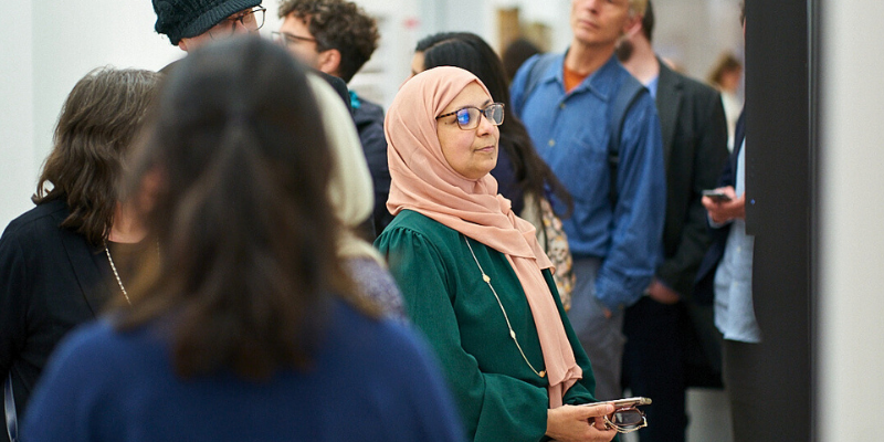 A woman wearing glasses and a headscarf looks at a work of art on a wall, surrounded by other gallery-goers