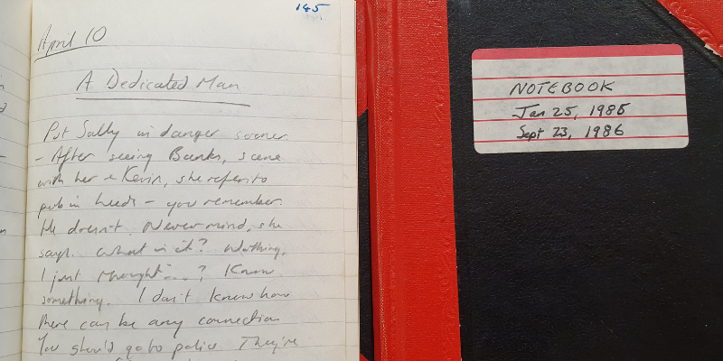 An open notebook with the words "A Dedicated Man" written at the top of the page alongside a closed notebook.