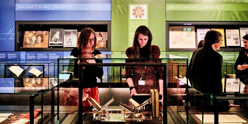 Two women look at a display case in the Treasures of the Brotherton Gallery, against a blue and green background.