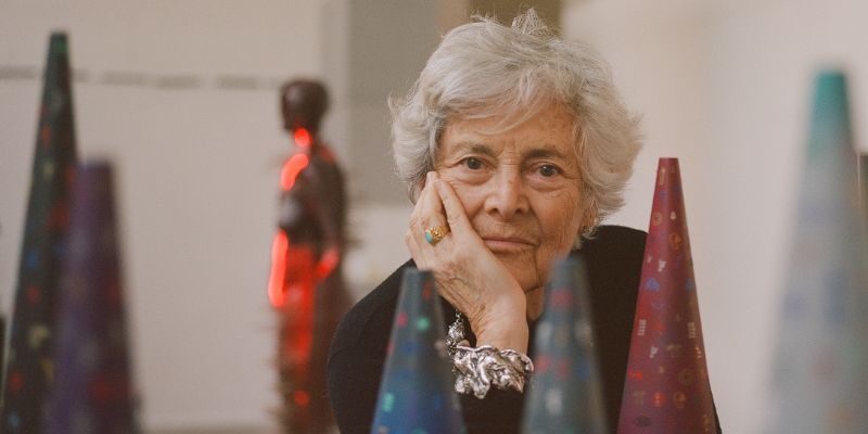 A woman with short grey hair looks at the camera with her head resting on her hand. Around her are cone shaped sculptures, some in and some out of focus.