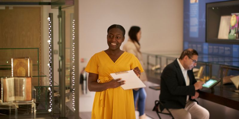 A person in a yellow dress holding a clipboard and writing in the middle of a museum space surrounded by glass cases. Behind them, two more people are looking at the displays.