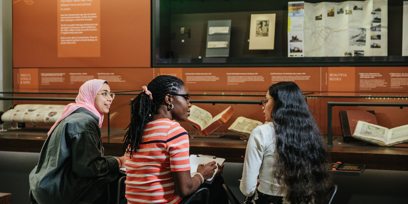 A woman stands in front of a sign saying 'Leeds'. She has a large book from Special Collections in front of her and she is presenting it to two students.