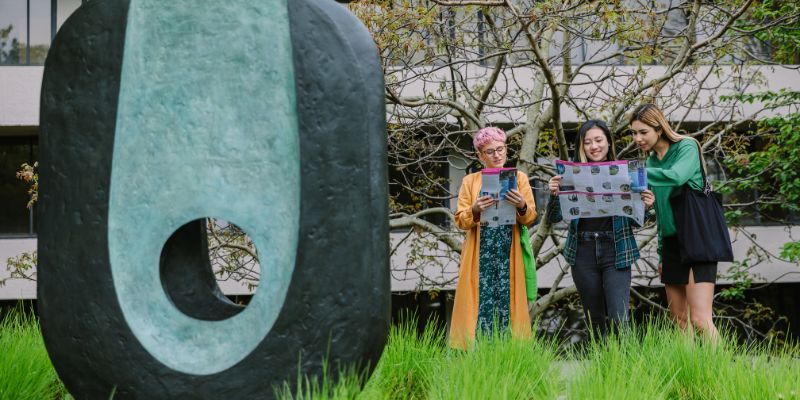 Three people looking at maps stood outside next to an oval shaped blue and black sculpture.