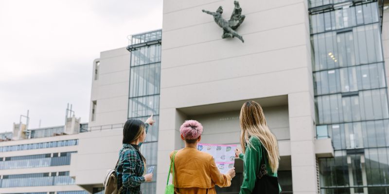 Three people looking at a map stood in front of a concrete building and pointing up at a sculpture on the wall.