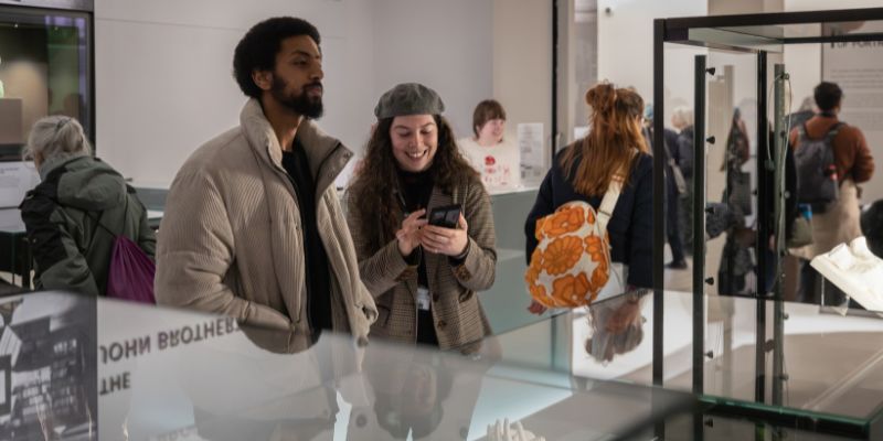 People stood in a museum surrounded by objects in glass cases. In the centre are two people stood together whilst one smiles at their phone.