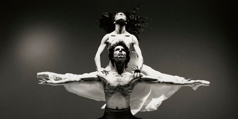 A black and white photograph of a topless male ballet dancer lifting a female dancer on his shoulders