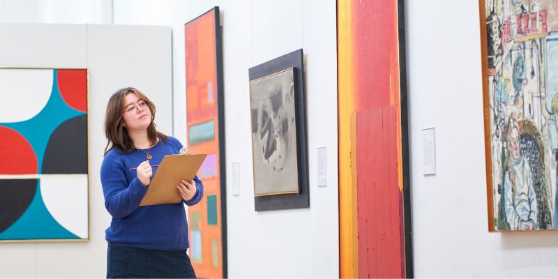 A group of students stood in The Stanley & Audrey Burton Gallery listening to a talk being given by a member of staff about one of the artworks on display.