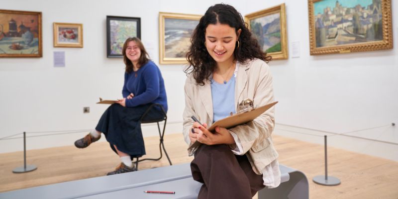 Two people sitting in a white gallery space surrounded by paintings and drawing on clipboards