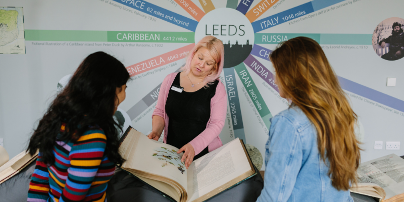 A woman stands in front of a sign saying 'Leeds'. She has a large book from Special Collections in front of her and she is presenting it to two students.