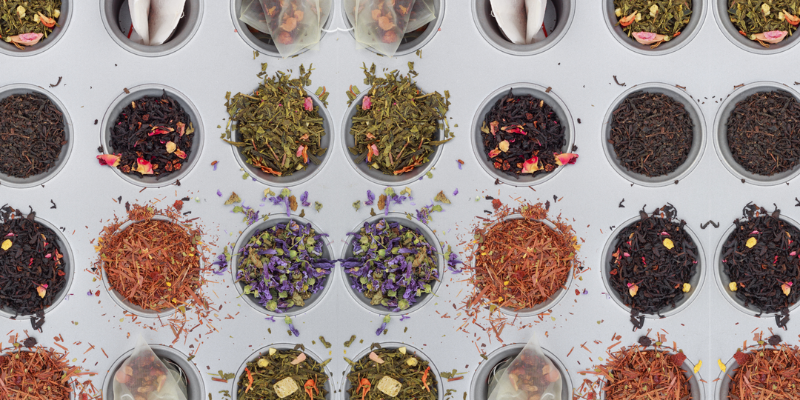 Photograph from above of rows of small round pots containing different coloured loose leaf tea blends on a grey background