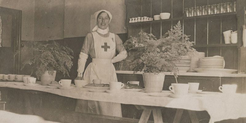 A back & white photograph of a woman in a nurse's uniform standing behind a trestle table laid with crockery and plants, smiling at the camera