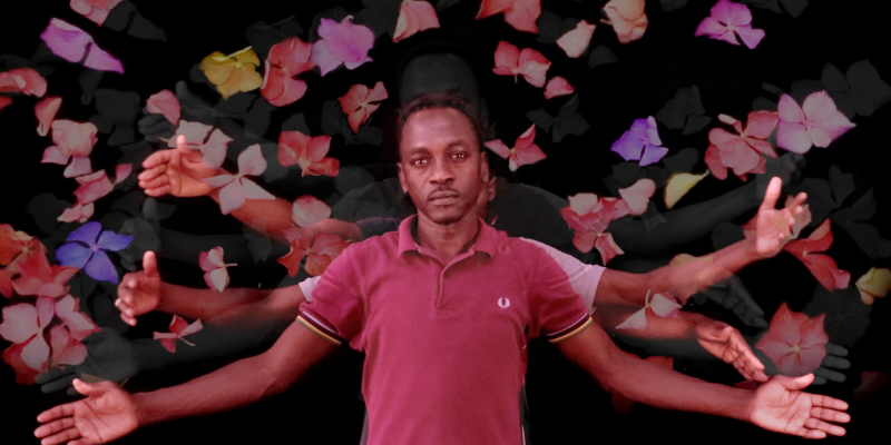 A man in a pink shirt with his hands outstretched, surrounded by flowers against a black background.
