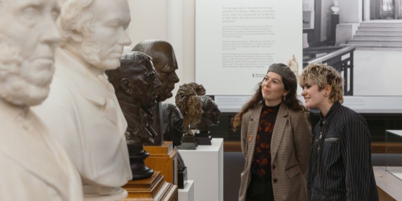Two people looking at a row of portrait busts made of bronze and marble, stood on plinths