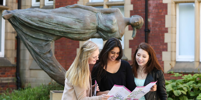 3 women holding a map, standing outside in front of a sculpture of a levitating women