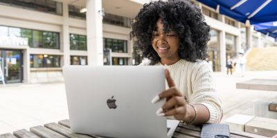 A smiling student is sat at a picnic bench using a laptop.