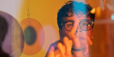 A young man wearing glasses is seen through a number of coloured glass lenses, with blue and orange light over his face. He raises his hand in front of his face to adjust one of the lenses.