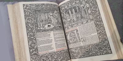 Open book on a grey cushion. The pages of the book are heavily decorated in black and white floral illustrations resembling medieval designs.