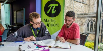 A student is sat at a table with a learning advisor showing them a book while talking. Behind them is a green banner with a value of pi symbol inside a black circle. Text underneath reads 'Maths Support'.