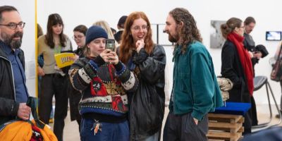 A group of people stood surrounded by art in a white walled gallery. They are smiling and one is taking photos on a mobile phone.