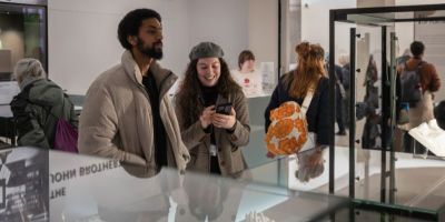 People stood in a museum surrounded by objects in glass cases. In the centre are two people stood together whilst one smiles at their phone.
