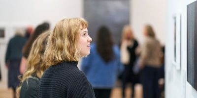 A white woman with short blond hair looks at an art work on a gallery wall. In the background are many other people exploring the exhibition.