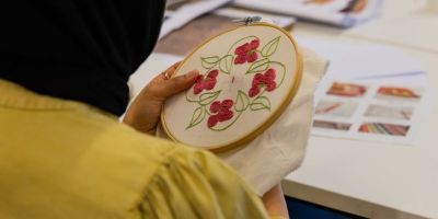 A person in a yellow top and black hijab holding an embroidery hoop and sewing a floral pattern.