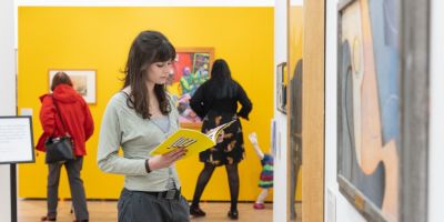A photo of someone with long black hair wearing a pale green cardigan looking at a yellow catalogue whilst stood in an exhibition. The wall behind is painted yellow.