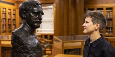 A person with short brown hair and wearing a black shirt looks at a bronze bust of a man with a moustache on a wooden plinth. They are in a wood-panelled room.