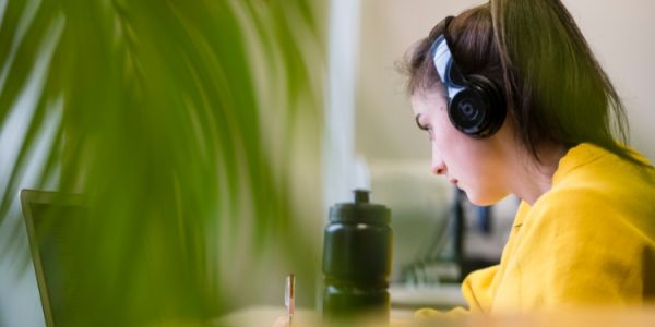 Behind a plant is a student at a desk. They're wearing headphones and looking at their laptop.