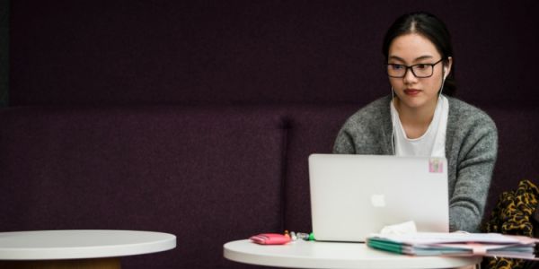 A student is studying at a table with a laptop. The seating and wall behind them is a deep purple.