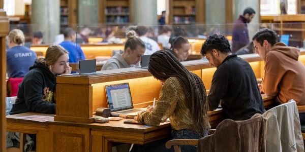 Students sit working with laptops at a long oak desk with task lighting in a busy Brotherton reading room.