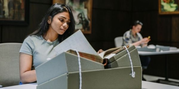 A woman is sat at a table with an open book on a book stand. She is flipping through the pages.