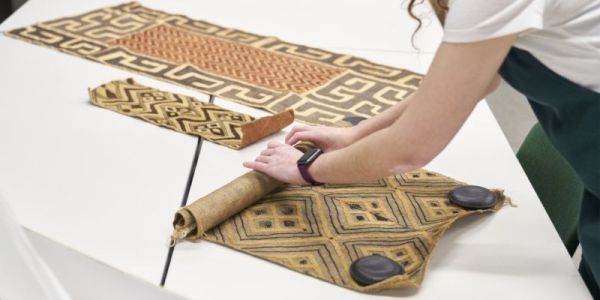 Three rectangular pieces of fabric tapestry are on a white table. A person is rolling one of the pieces.