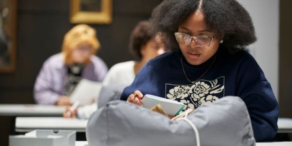 A person is seated at a table. They examine an artefact on a grey cushion and write in a notebook.