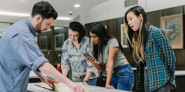 Four people stand around a table looking at an old book. One takes a photo with a phone.