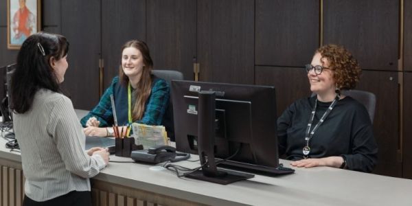 Two people sit smiling behind a reception desk with a monitor on it. Another person stands infront of the desk, engaging in conversation.