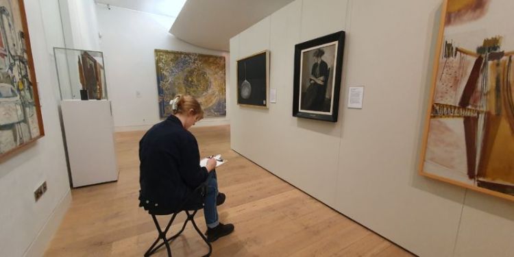 Isabelle, a white woman with blonde hair, sitting on a gallery stool with a clipboard and pencil drawing paintings in The Stanley &amp; Audrey Burton Gallery.