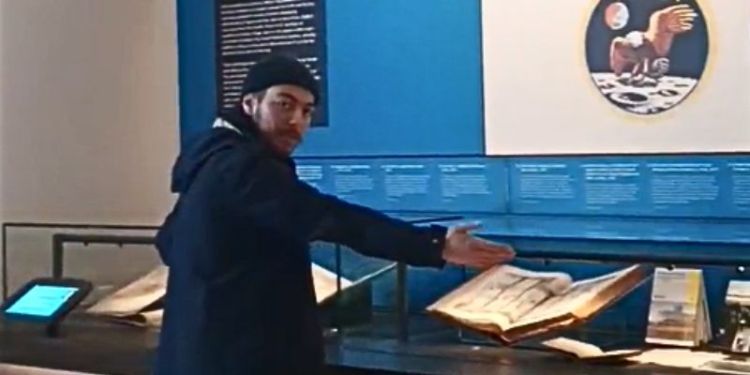 Matthew, a white man with dark hair wearing a navy coat, pointing at objects in glass cases in Treasures of the Brotherton