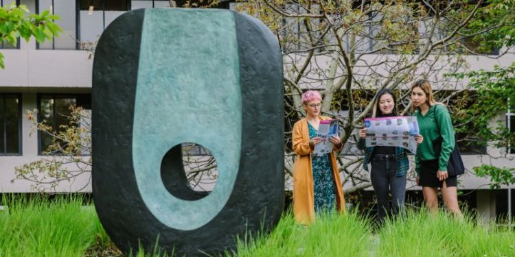 Three people holding public art maps beside a blue and black oval sculpture with a hole through it