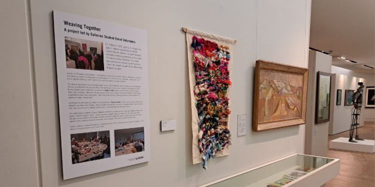 A text panel with the title "Weaving Together" beside a colourful hanging weaving and a painting of an abstract landscape in oranges and browns, all on a white gallery wall.